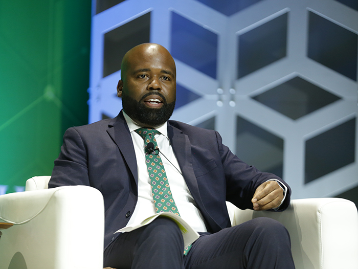 A person in a suit and tie sits in a white cozy chair on a stage and talks to the audience. 