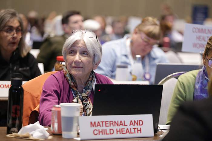 A woman with white hair and a pink blazer sits in front of a laptop and a sign that says Maternal and Child Health. 