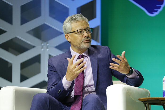 A man in a suit and purple tie sits in a white cozy chair on a stage and talks with his hands while looking toward someone off camera. 