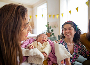 A mother holds her infant as a woman looks on smiling, both seated on a couch