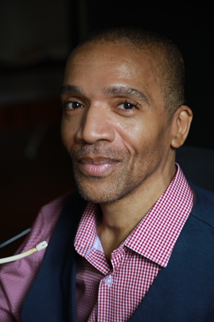 Black man in a red and white checkered collared shirt and jacket, looking at the camera with a slight smile