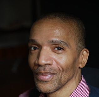 Black man in a red and white checkered collared shirt and jacket, looking at the camera with a slight smile