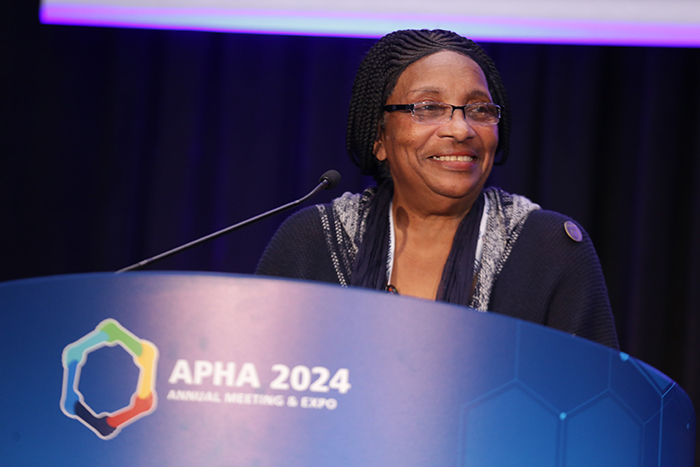 APHA President Ella Greene-Moton stands at a lectern on a stage and smiles at the crowd. 