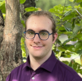 White person smiling at the camera wearing glasses and a purple collared shirt, standing outside