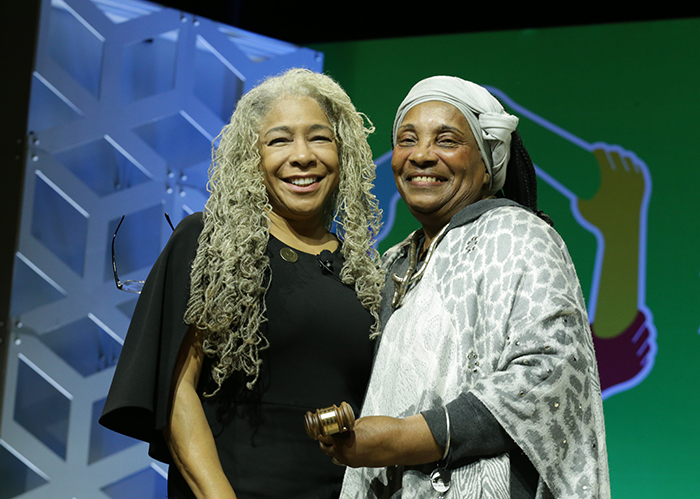 Two Black women stand close together on a stage and smile for the camera. 