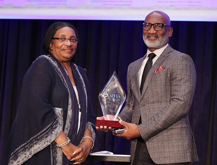 Two people stand next to each other and pose for the camera while one of them holds an award. 