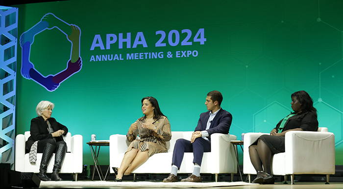 Four people sit on white chairs on a big stage, with everyone looking at the the woman speaking. 