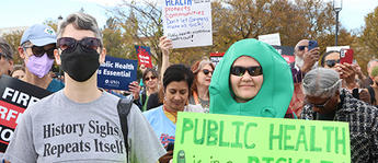 A woman wearing a black face mask wears a T-shirt that says "History sighs, repeats itself. Next to her is a woman wearing a pickle costume holding a sign that says "public health is in a pickle."
