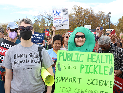 A woman wearing a black face mask wears a T-shirt that says "History sighs, repeats itself. Next to her is a woman wearing a pickle costume holding a sign that says "public health is in a pickle."