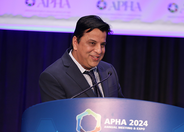 A person stands at a lectern with the APHA Annual Meeting logo on it while smiling at the crowd. 