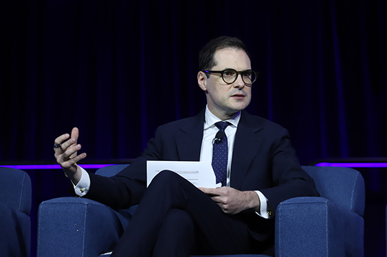 Richard Hughes, dressed in a dark suit and tie and holding a sheet of paper, sits on a comfy blue chair on a stage and talks to the audience. 