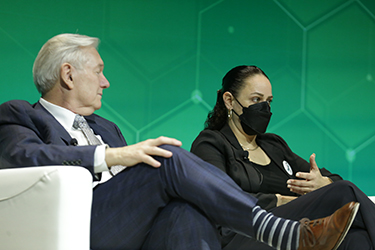 Michael Osterholm and Karyn Bishof sit on white chairs on a stage while Karyn, wearing a black face masks, talks to the audience and uses her hands. 