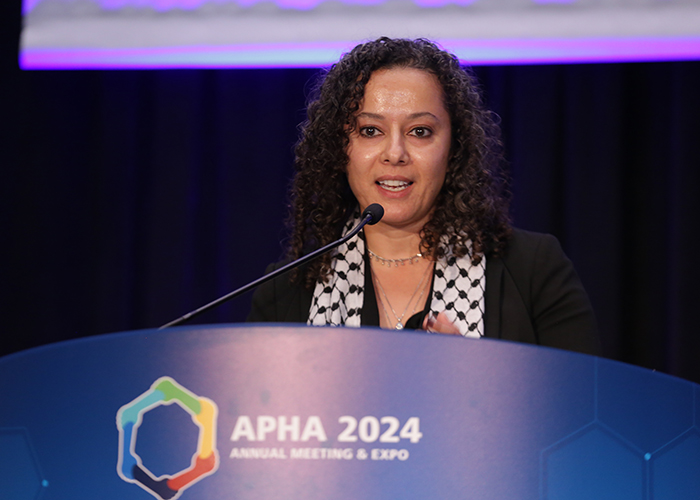 A person stands at a lectern with the APHA Annual Meeting logo on it while speaking into the microphone. 