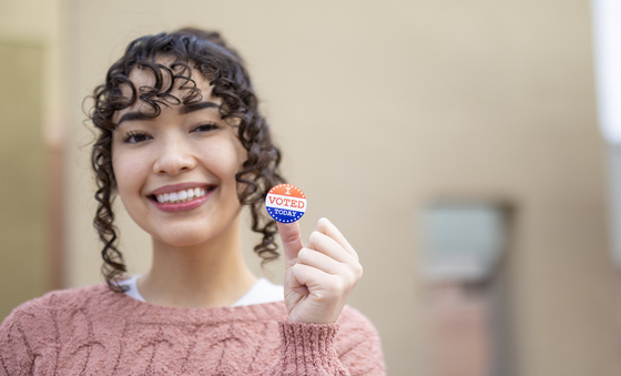 Woman with brown curly hair in a pink sweater smiling and holding an I Voted sticker