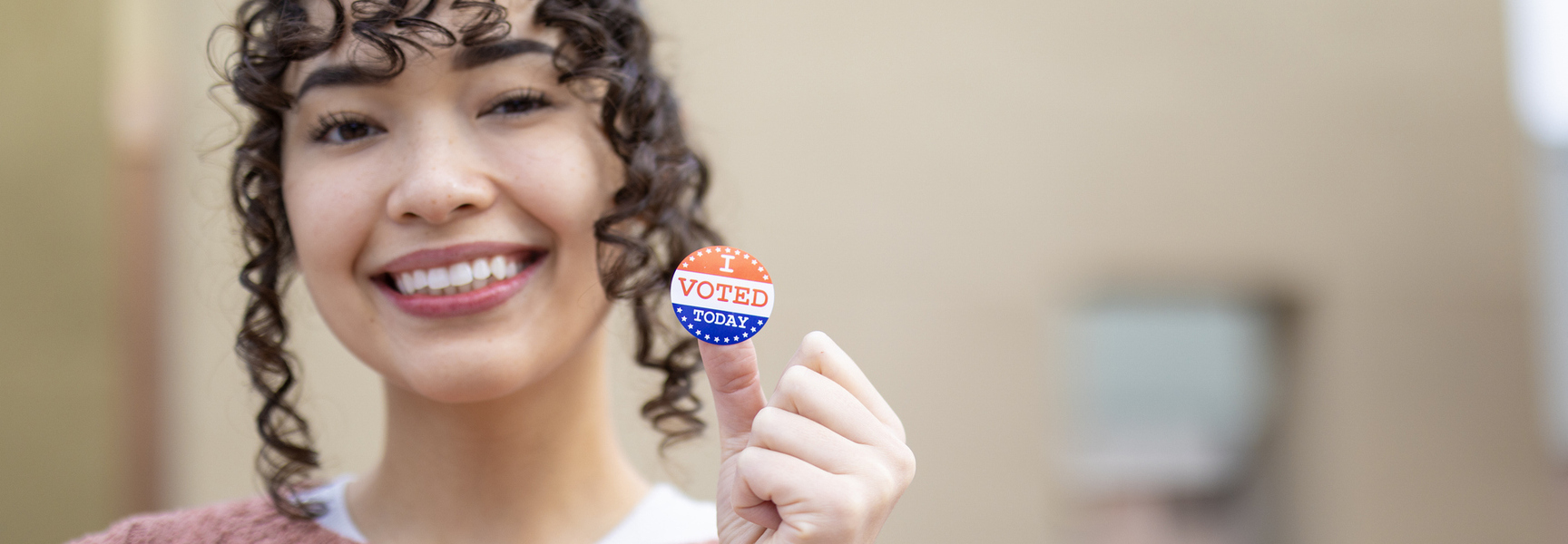 Woman with brown curly hair in a pink sweater smiling and holding an I Voted sticker