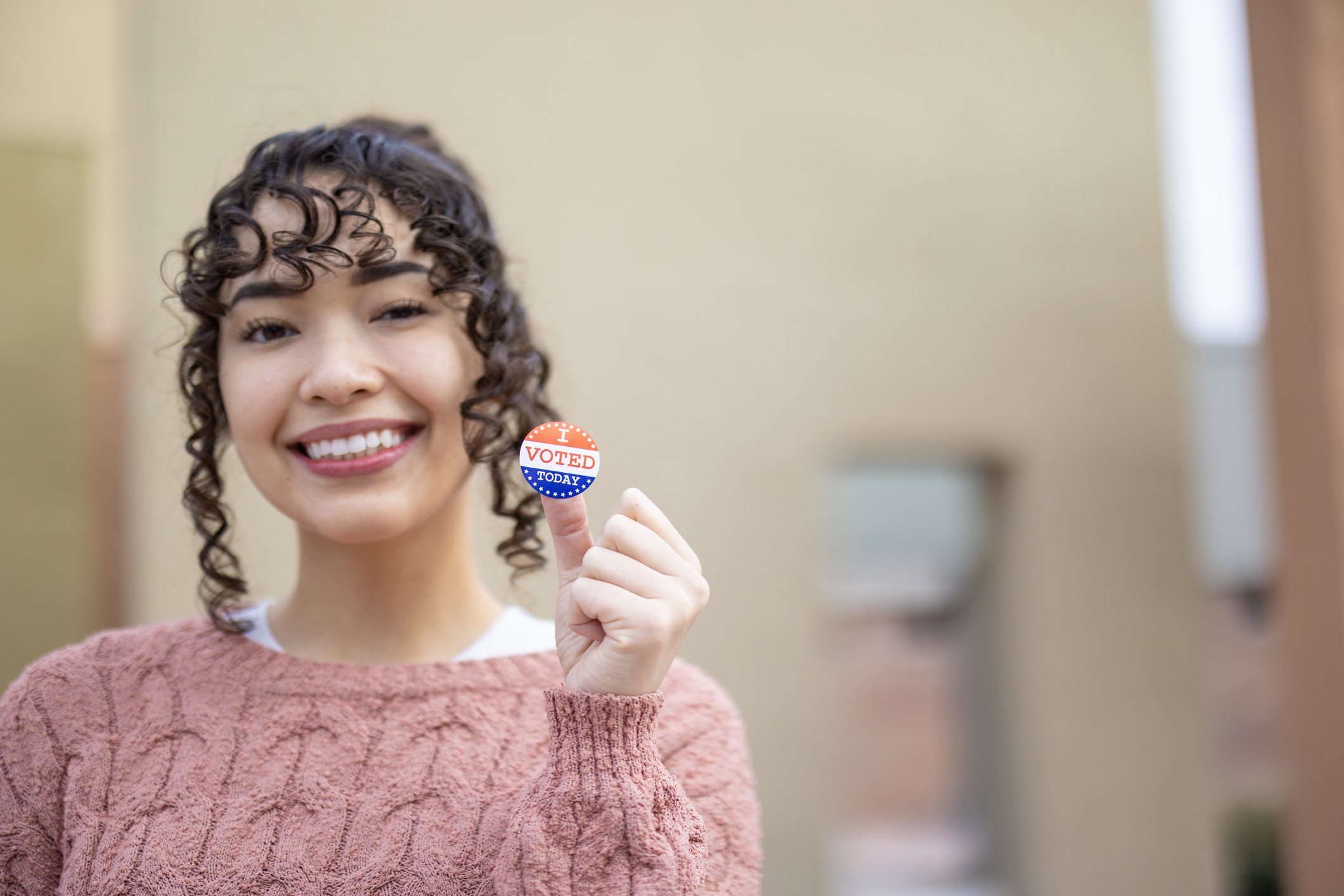 Woman with brown curly hair in a pink sweater smiling and holding an I Voted sticker