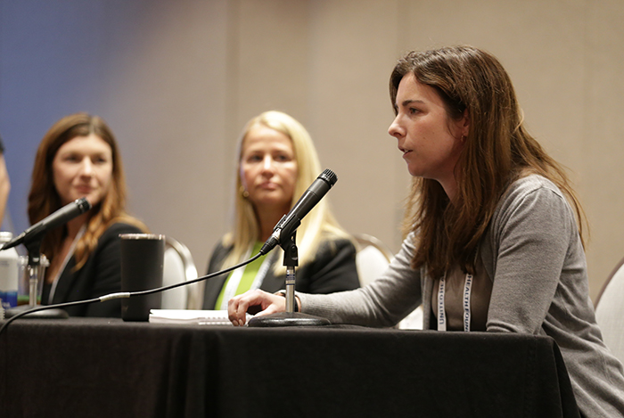 Three women on a panel sit at a table with microphones and one woman is talking. 