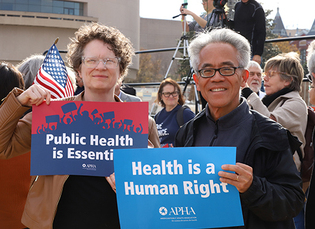 A man and woman stand next to each other at a rally outside. She holds a "Public health is essential" sign and his sign says "health is a human right." 