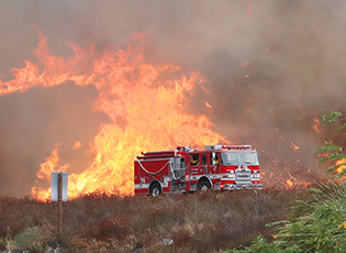 A firetruck sits in the foreground of a wildfire sweep across hills