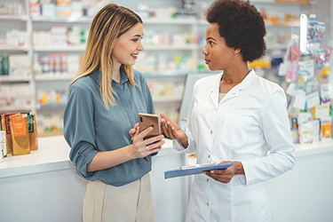 A customer shows a pharmacist something on her phone while they chat in front of a pharmacy counter. 