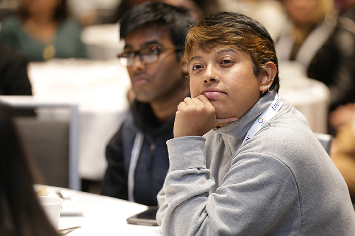 A young person sits at a table with their head resting on their fist and elbow resting on the table as they look at a presenter. 