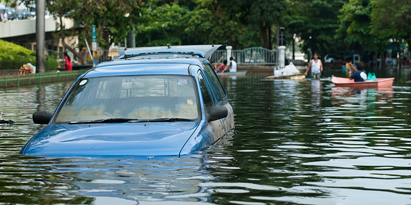 Blue car stuck in a flooded street with water above the headlights and ple in the background