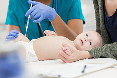 A white baby lays on a table while a woman holds its hands and a person in scrubs and wearing blue latex gloves gives a shot in the baby's thigh. 