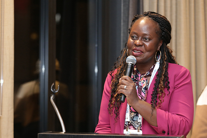 Black woman in bright pink jacket stands at a lectern and holds a hand microphone in her left hand while speaking and smiling 