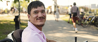 A man looks at the camera smiling while sitting on a power mobility device in a park