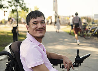 A man looks at the camera smiling while sitting on a power mobility device in a park