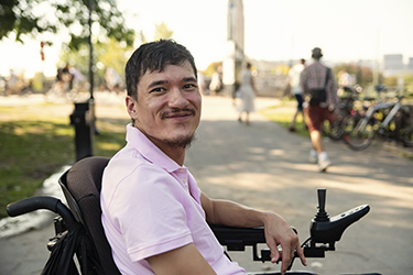 A man looks at the camera smiling while sitting on a power mobility device in a park