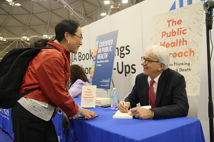 A woman in maroon casual jacket and black backpack leans forward on a table talking to a male author signing a book for her, as the two talk. 