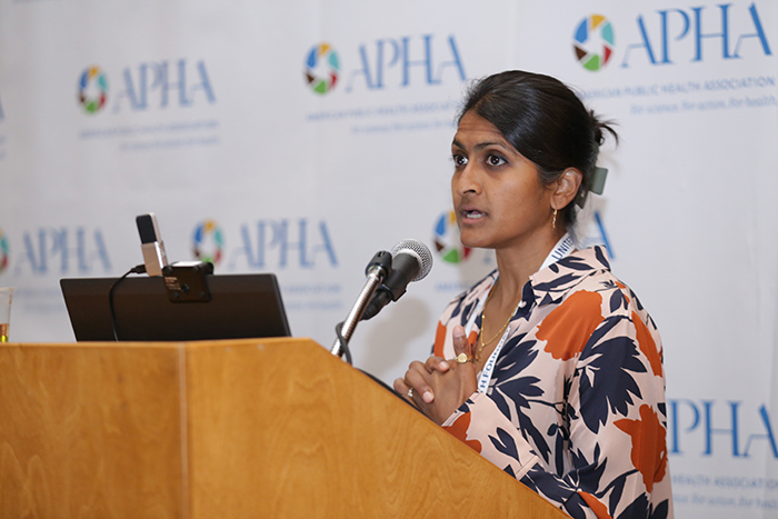 A woman in a blue and orange flowered shirt talks at a lectern with a microphone and laptop on it. 