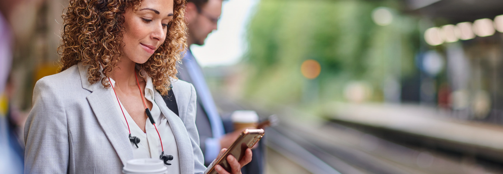Woman with curly brown hair dressed professionally with a coffee cup in one hand and her phone in the other waiting for the train