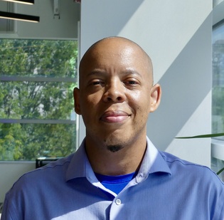 A Black person smiling at the camera wearing a blue collared shirt and lots of greenery in the background