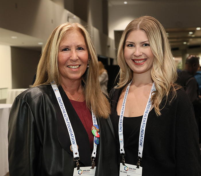 Two blonde women dressed in black business casual stand next to each other and smile at the camera. 