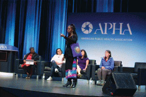 four seated people one woman standing on stage