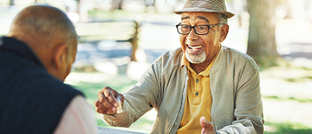 Two older Black men sit at an outdoor table and play chess. One is making a move and smiling, while we can only see the back of the other man. 
