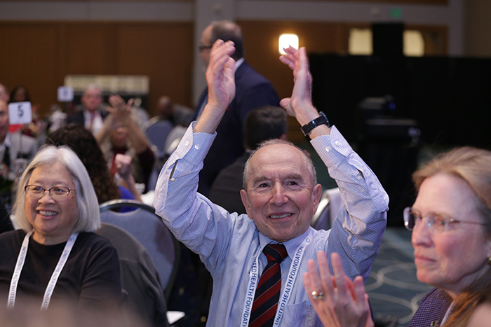 A man sitting at a table smiles and raises his hands above his head to clap. There are two people sitting on either side of him. 