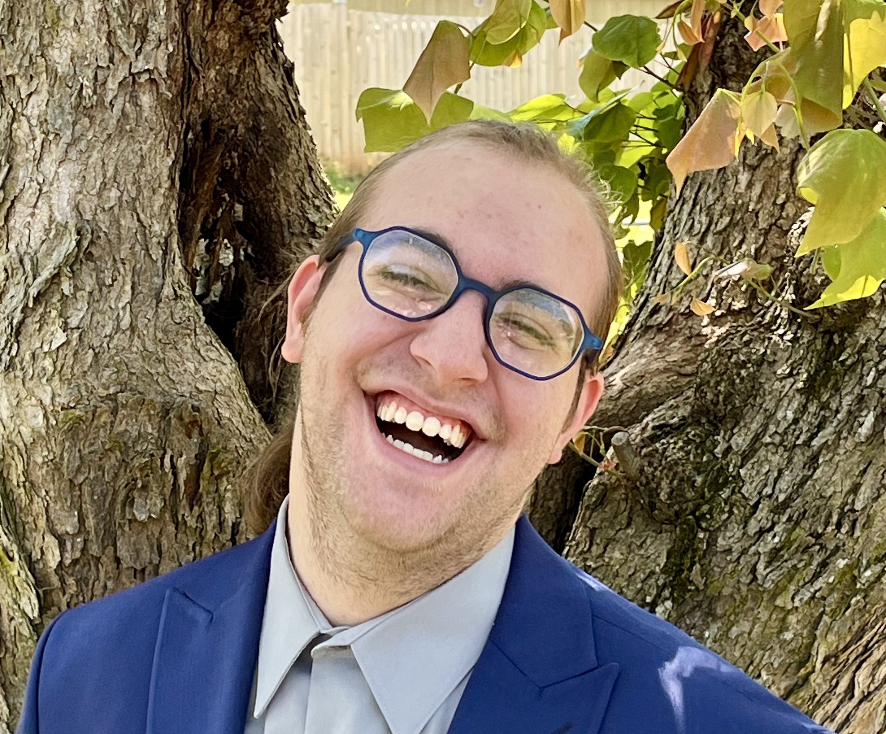 A person with glasses laughing with their head tilted to the side, they are standing in front of a tree 