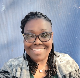 A Black woman with braids in front of a blue background, smiling with glasses on. 