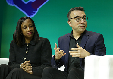 J. Nadine Gracia is dressed all in black and sits in a white chair with her hands clasped on her lap, while she watches Bechara Choucair next to her talk during a panel discussion. 
