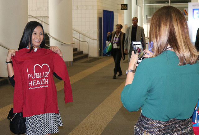 woman takes photo of another women holding I Love Public Health shirt