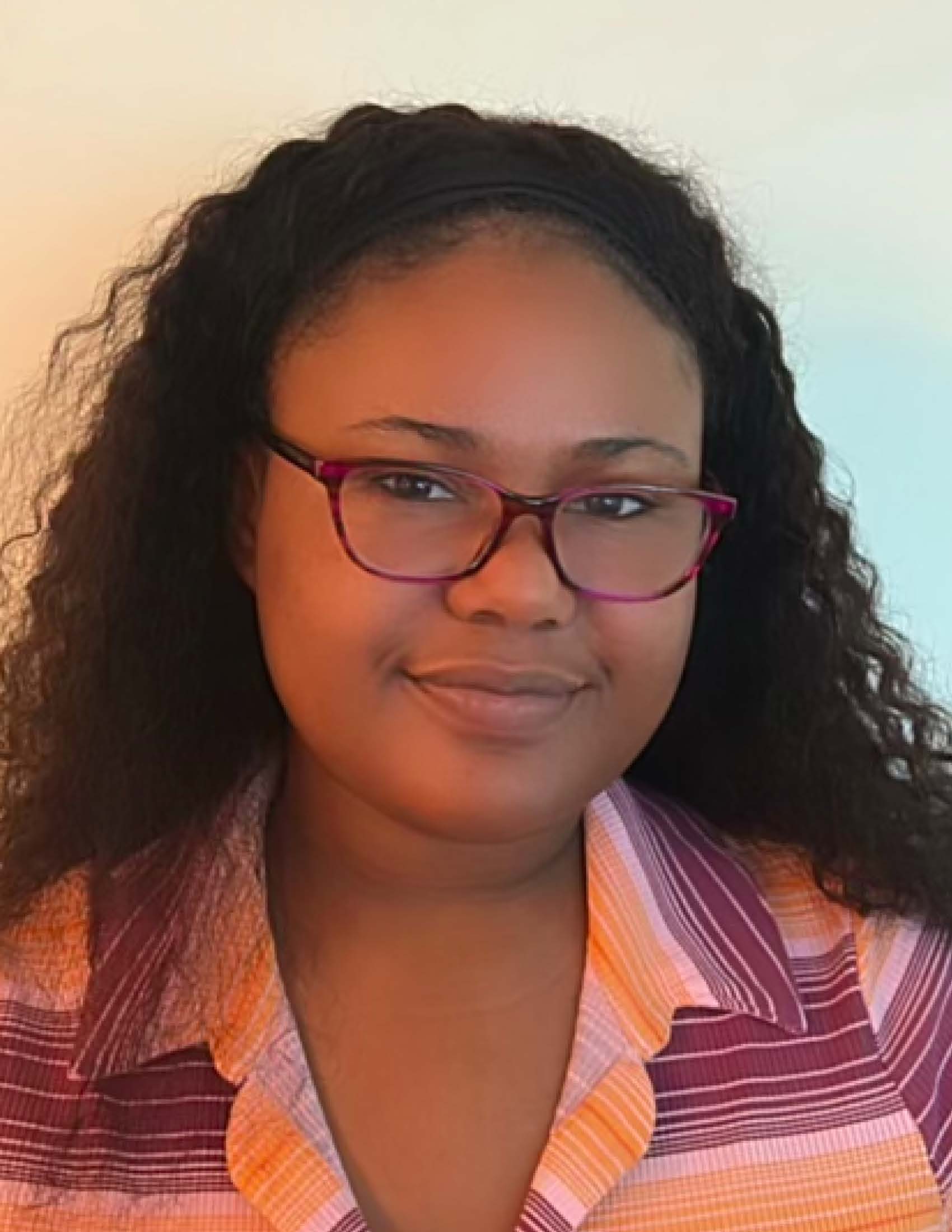 A Black person smiling at the camera with long dark hair and glasses, wearing a colorful collared shirt 