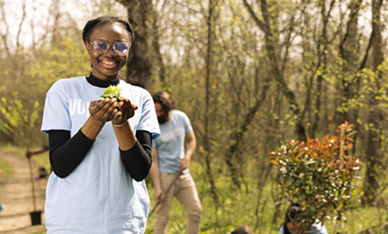 A Black teenager girl stands in nature and holds small plant in her hand while smiling at the camera while others plant a tree behind her. 