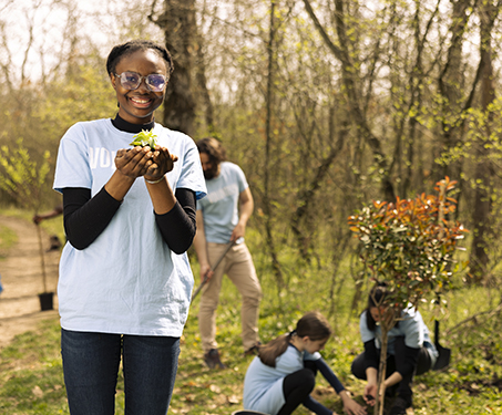 A Black teenager girl stands in nature and holds small plant in her hand while smiling at the camera while others plant a tree behind her. 