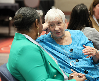 A Black woman and a white woman sit in chairs next to one another, facing each other and talking. 