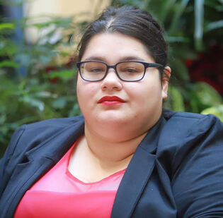 Latina woman in a suit and glasses wearing red lipstick with greenery in the background 