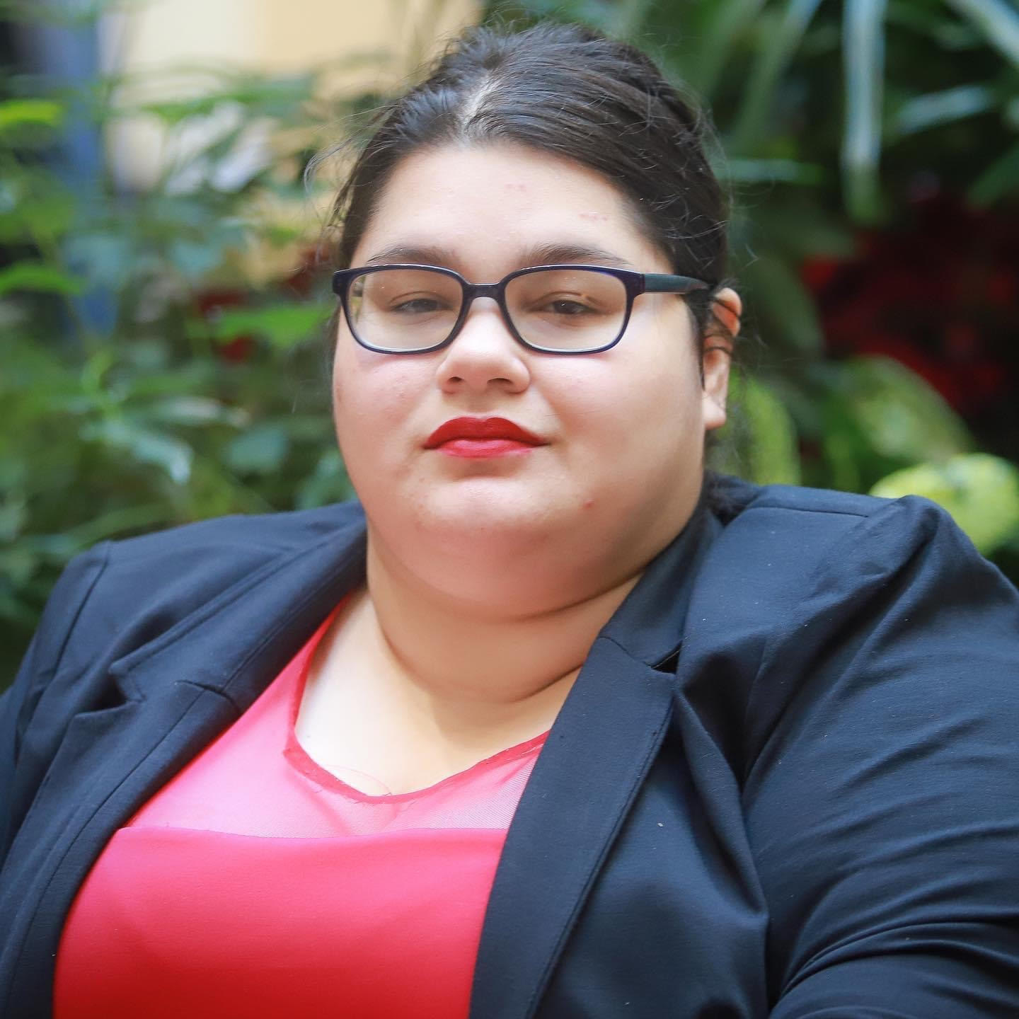 Latina woman in a suit and glasses wearing red lipstick with greenery in the background 