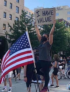 masked black woman at protest holding up I Have Value sign next to American flag